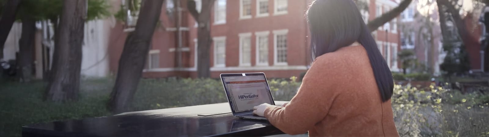 A student in an orange sweater works on a laptop at an outdoor table surrounded by trees and red brick buildings at the University of Florida campus.