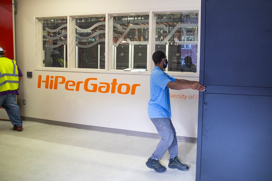A person in a blue shirt and mask moves equipment inside a facility, with a windowed wall revealing computer servers and “HiPerGator” written in orange on the wall.