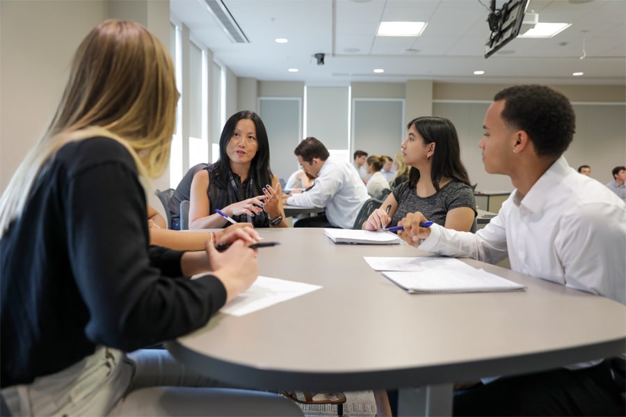 Four students sit together at a round table, actively discussing and taking notes as they collaborate on a project in a classroom setting.