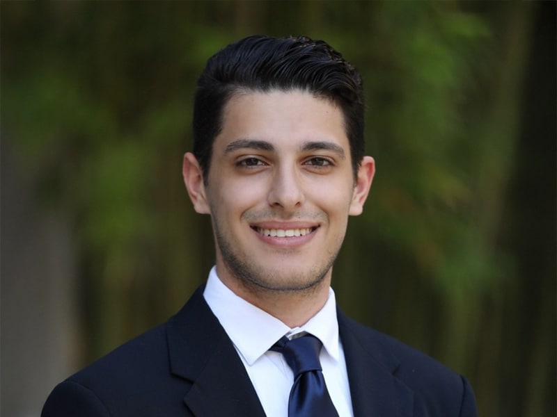 A young man with neatly styled dark hair and a confident smile poses in a navy suit and tie, standing in front of a lush green backdrop.
