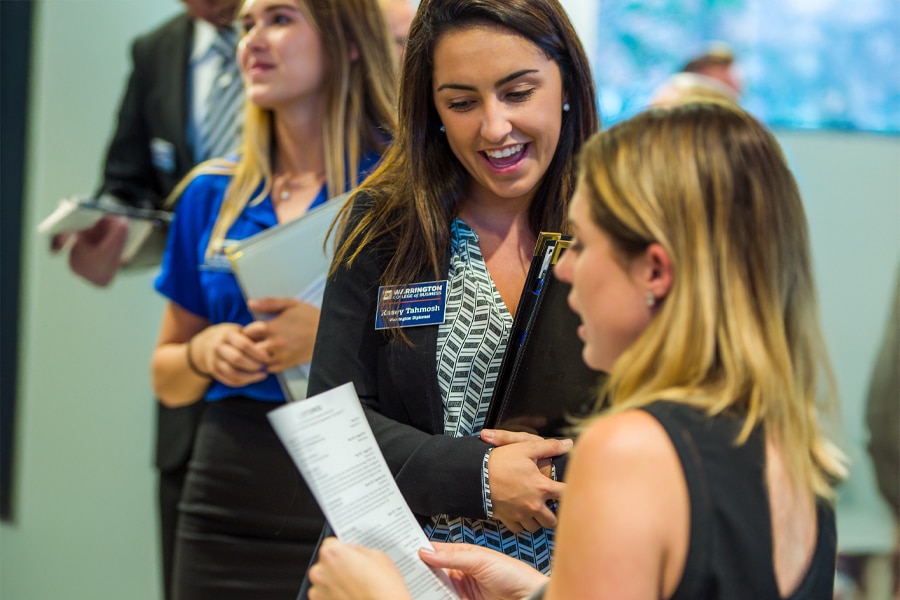 A Warrington College of Business student advisor named Kassy Talbmosh, holding a clipboard and smiling, talks with a student reviewing documents during an academic event.