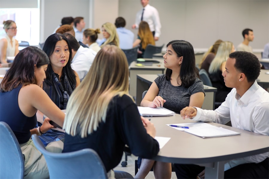 A diverse group of college students engages in a focused discussion around a table with an instructor guiding them; the classroom is busy with other groups in the background.