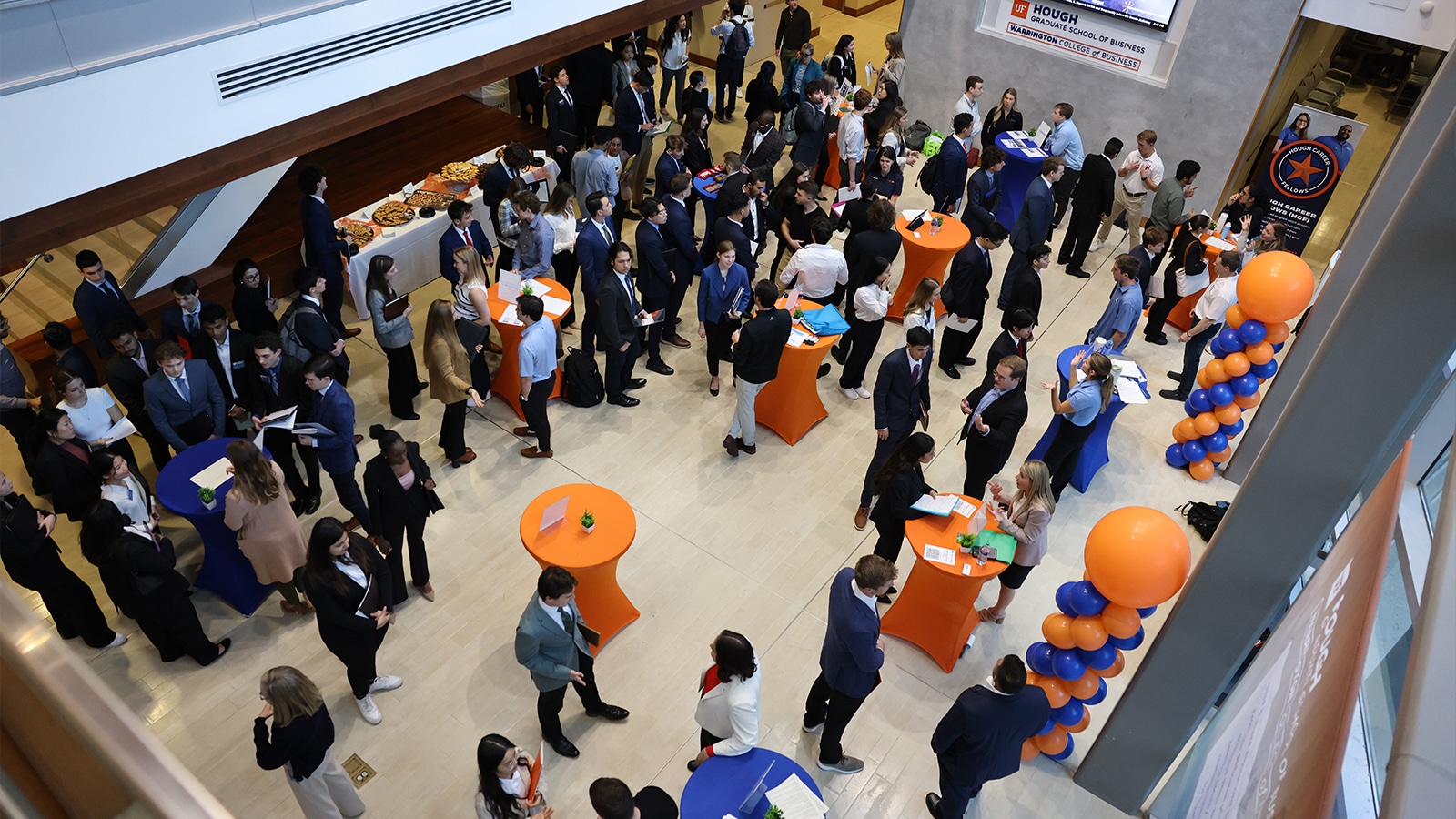 A large group of well-dressed graduate business students and professionals interact at a bustling networking event inside Hough Hall.