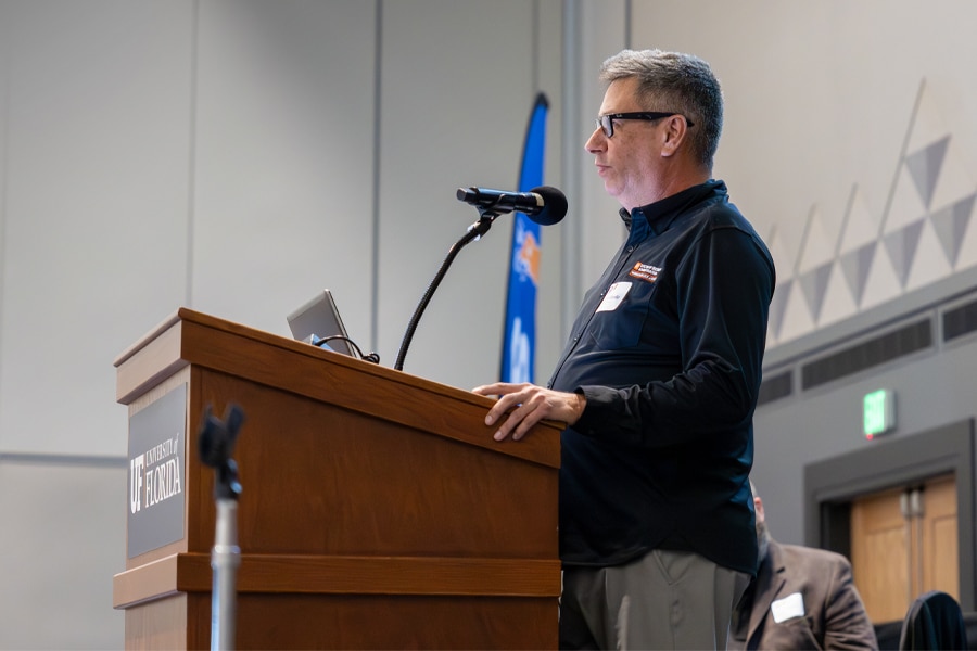 A male professor wearing glasses and a dark jacket speaks at a podium with a microphone in a large conference room at the University of Florida.