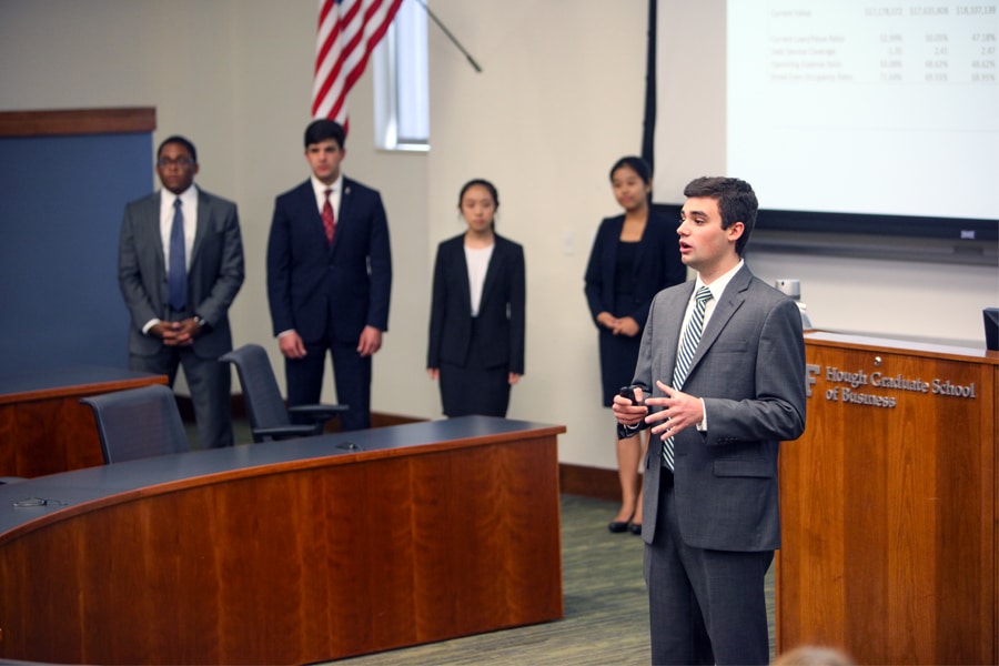 A business student in a suit delivers a presentation at the front of a classroom while four teammates stand ready in the background.