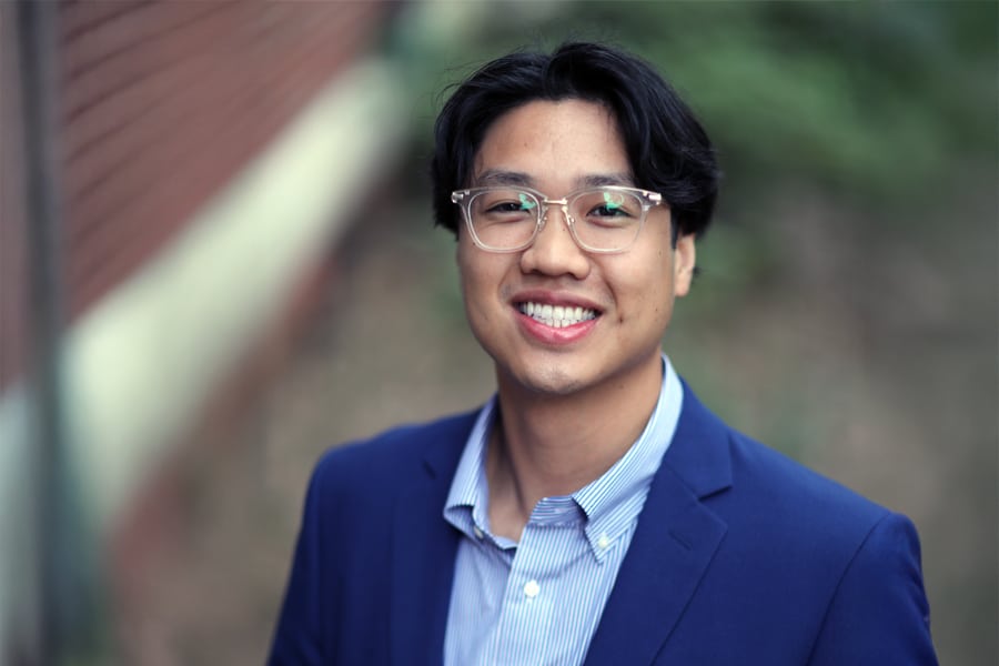 Portrait of Indy Lo, a young man with dark hair and glasses, wearing a blue blazer and a striped button-down shirt, smiling outdoors with a blurred background.