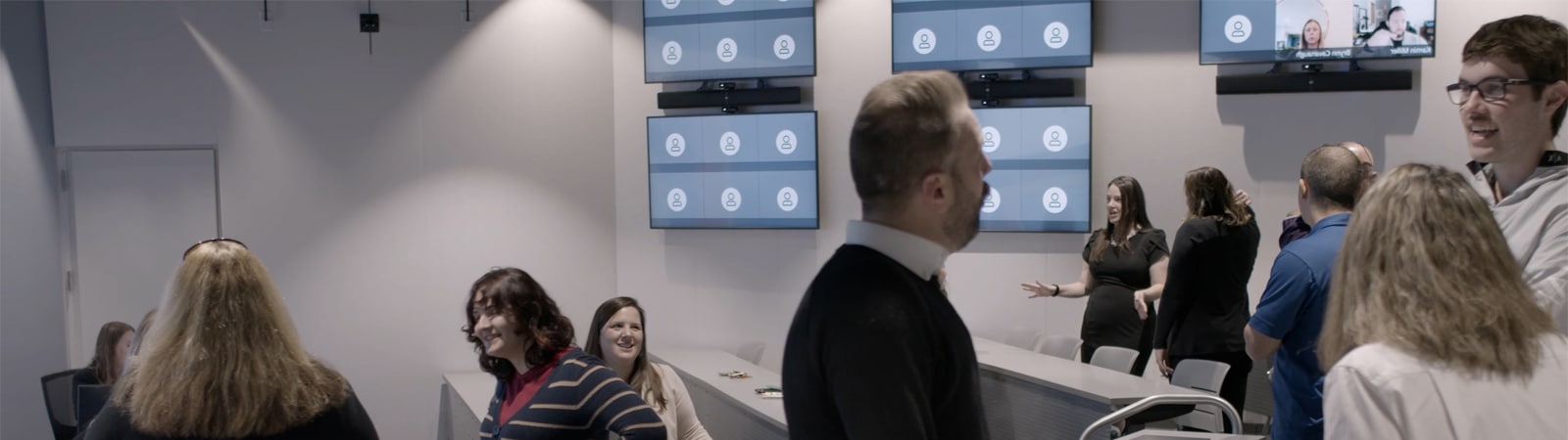 A diverse group of people interact and socialize in a modern classroom equipped with multiple screens displaying remote participants, blending in-person and virtual learning.