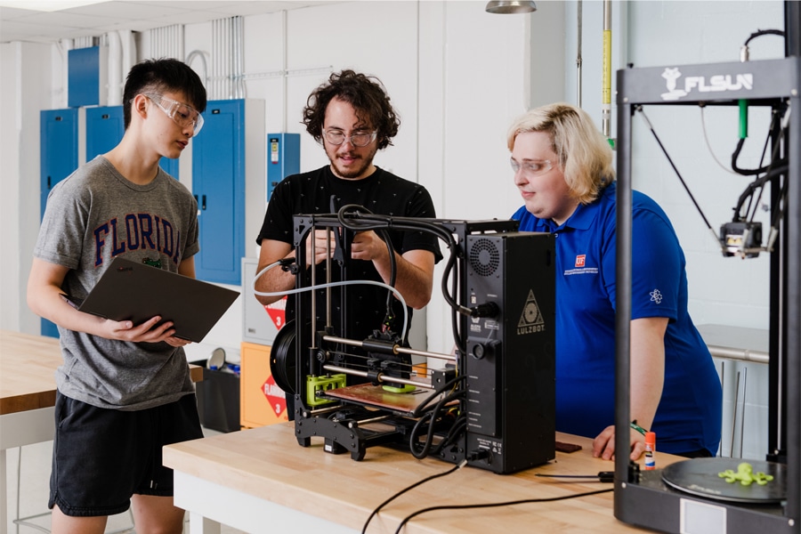 Three students wearing safety glasses collaborate on a 3D printing project in a workshop space. One is holding a laptop, another is working directly with the 3D printer, and a third is observing closely.