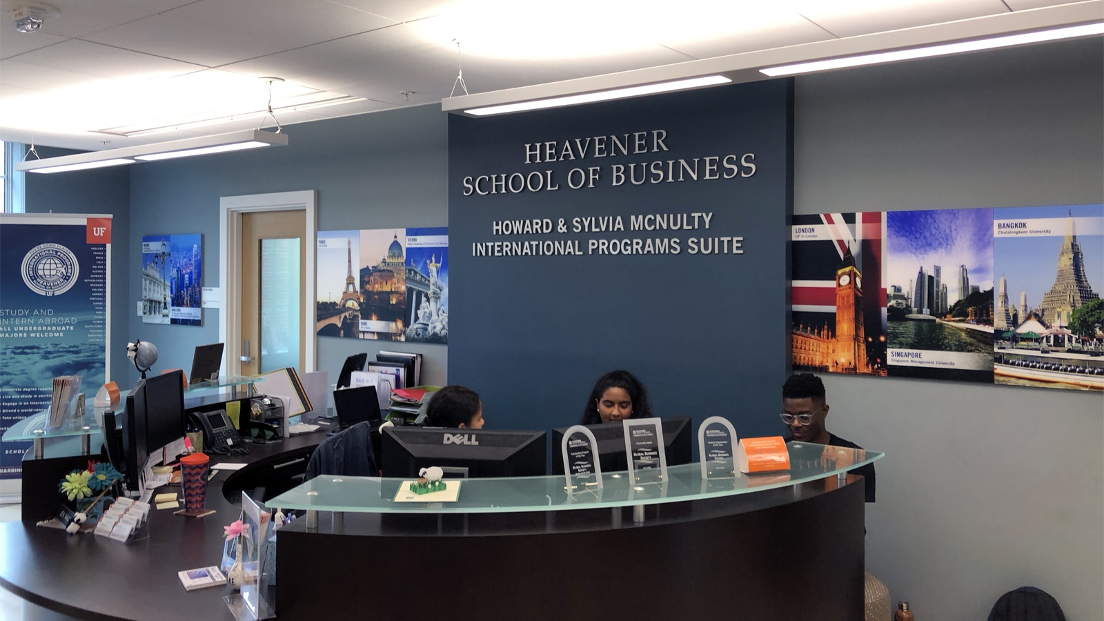 The reception area of the Howard & Sylvia McNulty International Programs Suite at the Heavener School of Business, showing a curved desk with staff members working behind it. Walls feature colorful travel posters from cities like Paris, London, Bangkok, and Singapore.