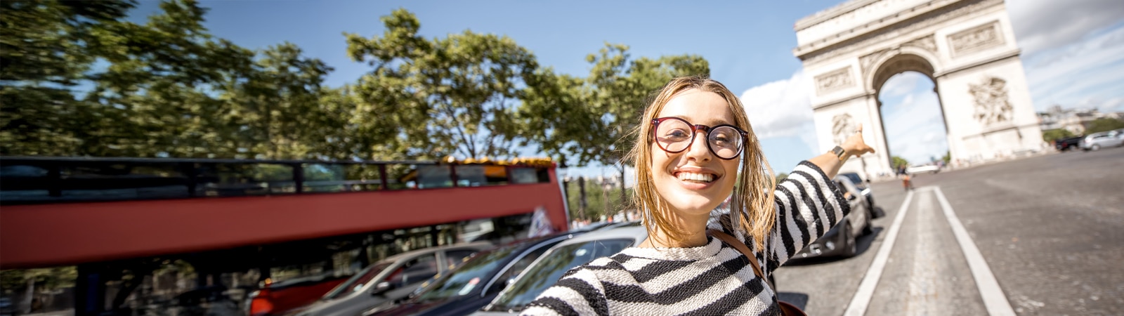 Smiling young woman wearing glasses and a black-and-white striped sweater points excitedly toward the Arc de Triomphe in Paris on a sunny day with blue skies and leafy green trees lining the street.