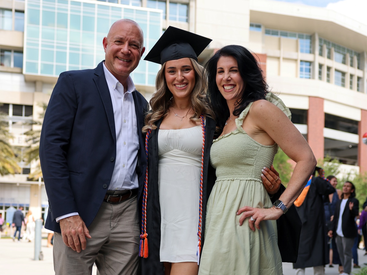 A female graduate in cap and gown stands between her parents outside a campus building on Commencement day.
