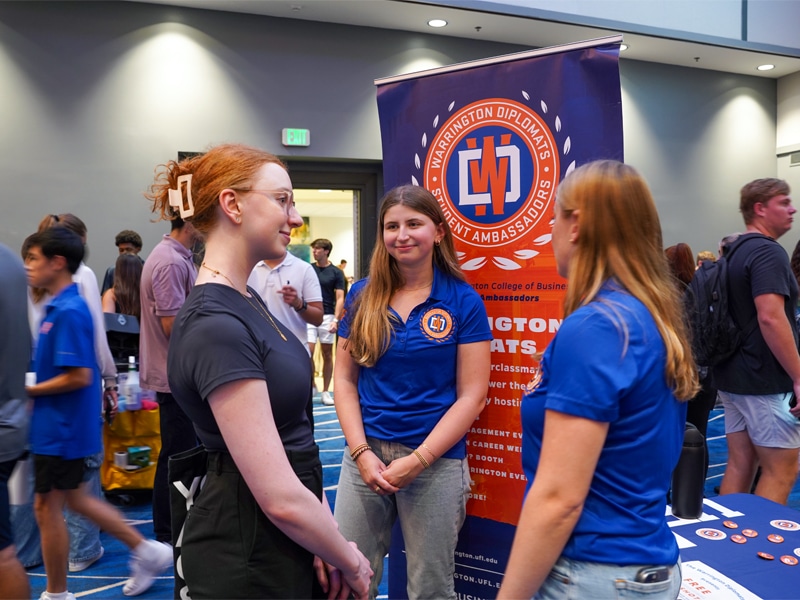 A student with red hair and glasses listens to two Warrington Diplomats — women in blue shirts with the Diplomats logo — at a busy, well-lit event. A colorful banner behind them highlights the Warrington Diplomats Student Ambassadors.