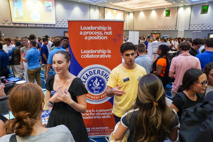 Students engage in conversation at a busy indoor event, standing near a banner that reads “Leadership is a process, not a position” and promotes the Leadership Development Program.