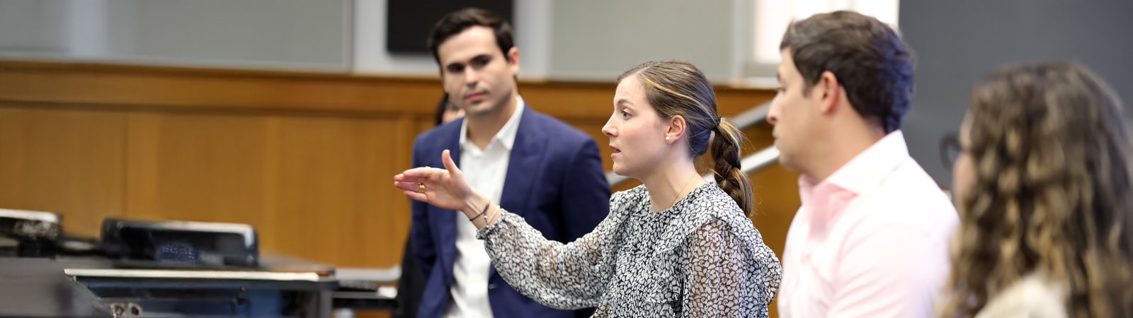 A woman in a patterned blouse speaks and gestures thoughtfully during a classroom discussion, while colleagues listen attentively in a professional setting.