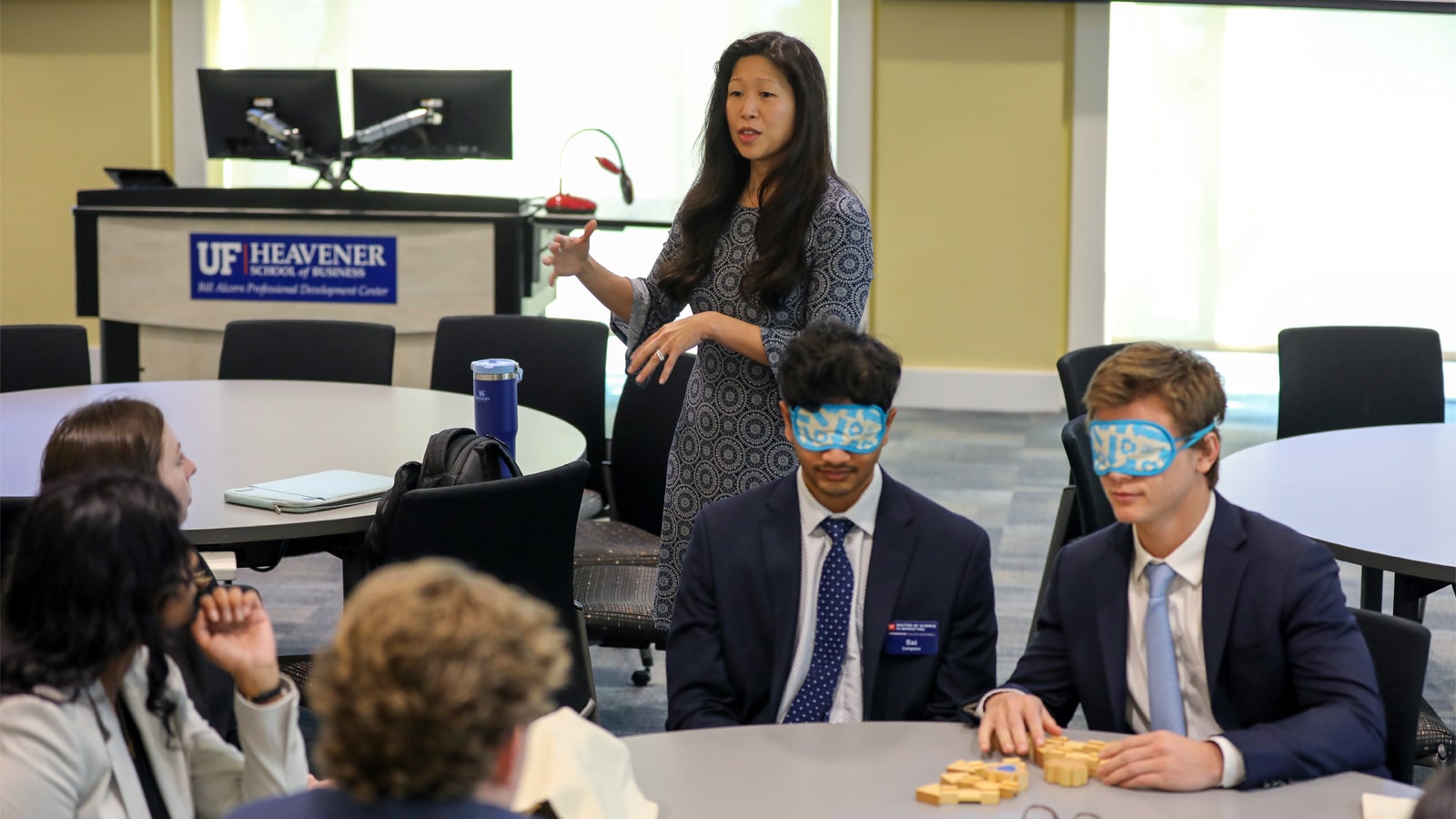 A facilitator leads an interactive activity with business students, two of whom wear blindfolds and sit at a table with wooden blocks in front of them, in a UF Heavener School of Business classroom.