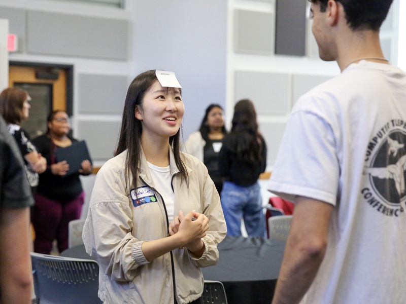 A smiling student with a name tag on her jacket and a sticky note on her forehead interacts with a peer during a group activity in a classroom. Other students converse in the background.