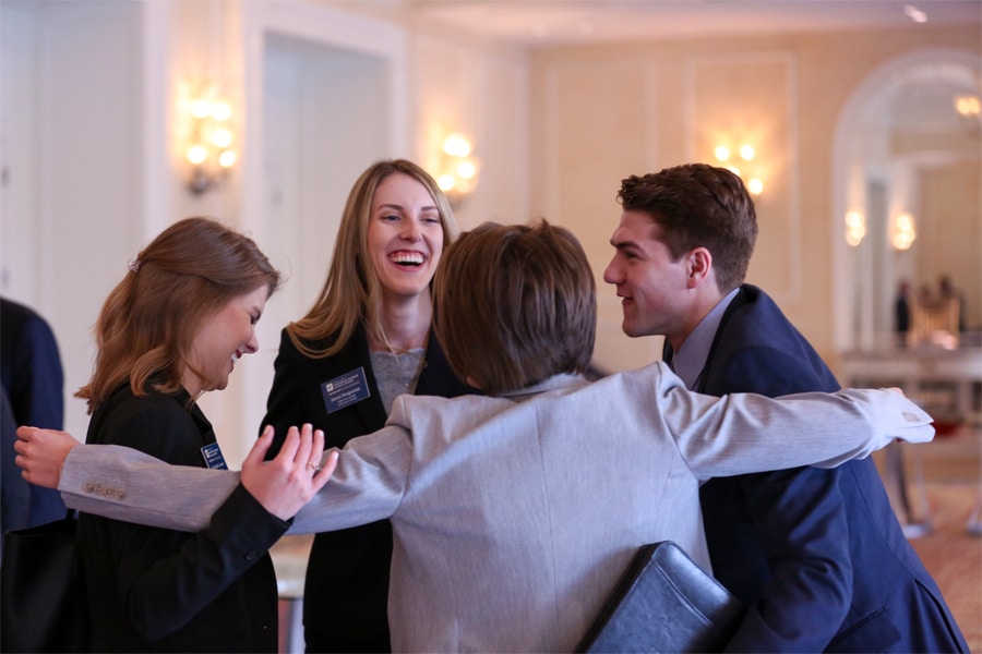 Four young professionals in business attire warmly embrace in a group hug while smiling and laughing in an elegant room.