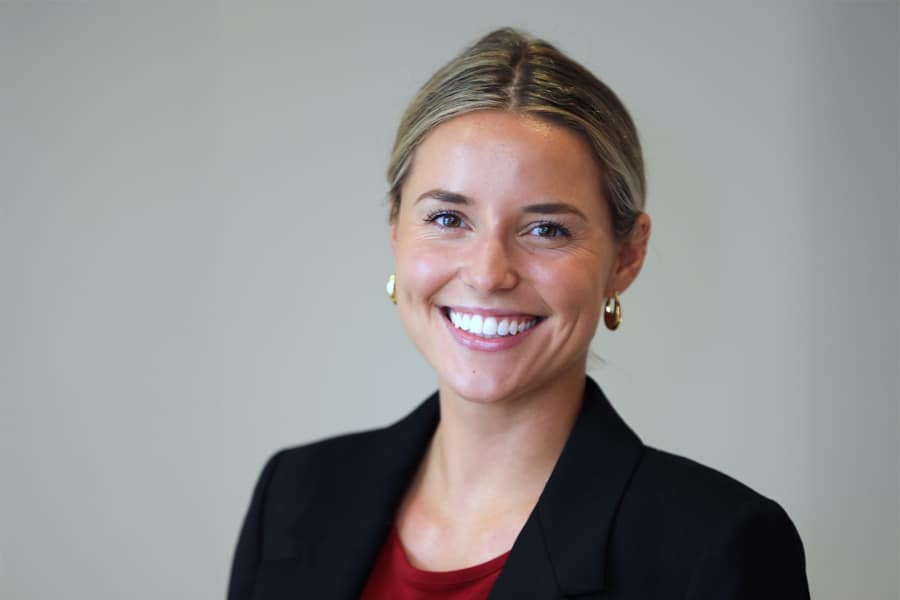 A young woman with blonde hair pulled back and gold hoop earrings smiles warmly at the camera, wearing a black blazer and red top, against a simple indoor background.