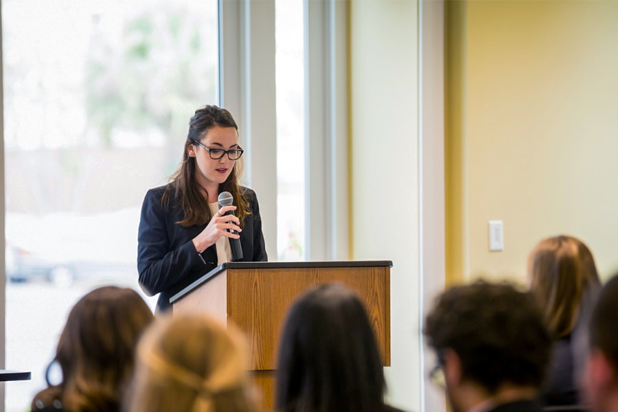 A young woman in professional attire speaks into a microphone at a podium in front of an audience of peers.