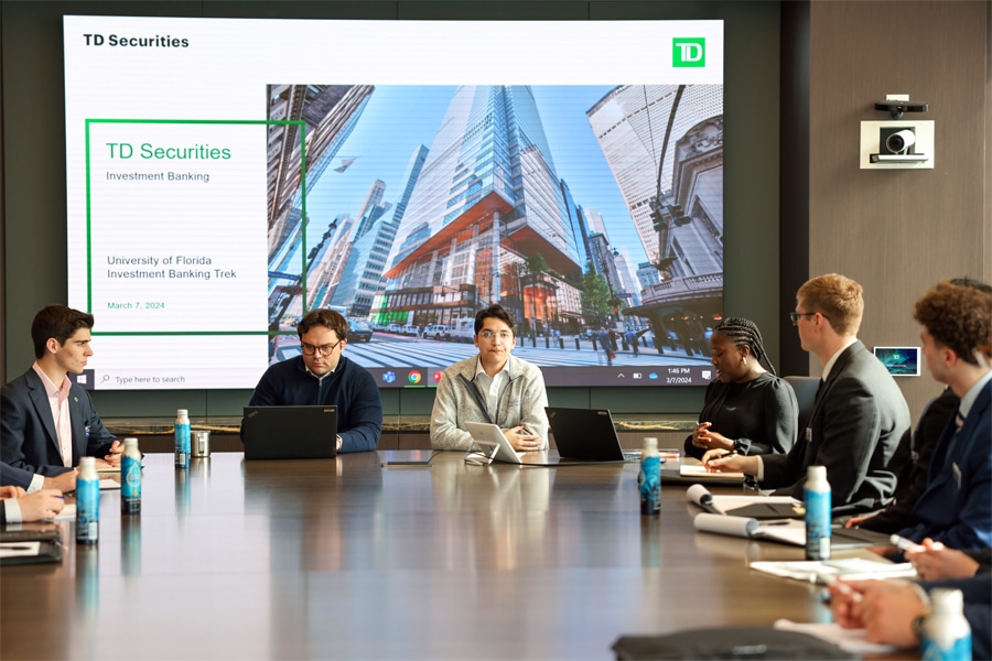 Students in business attire sit around a large conference table, collaborating with laptops and notebooks, while a presentation titled "TD Securities Investment Banking" is displayed on a screen behind them.