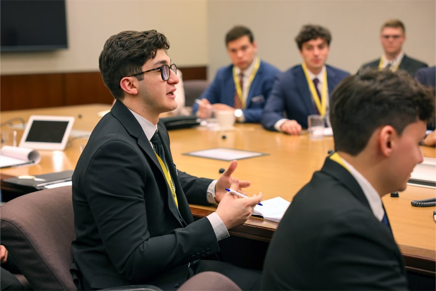 Business student in a suit participates in a boardroom discussion, gesturing as he speaks while others listen attentively around the table.
