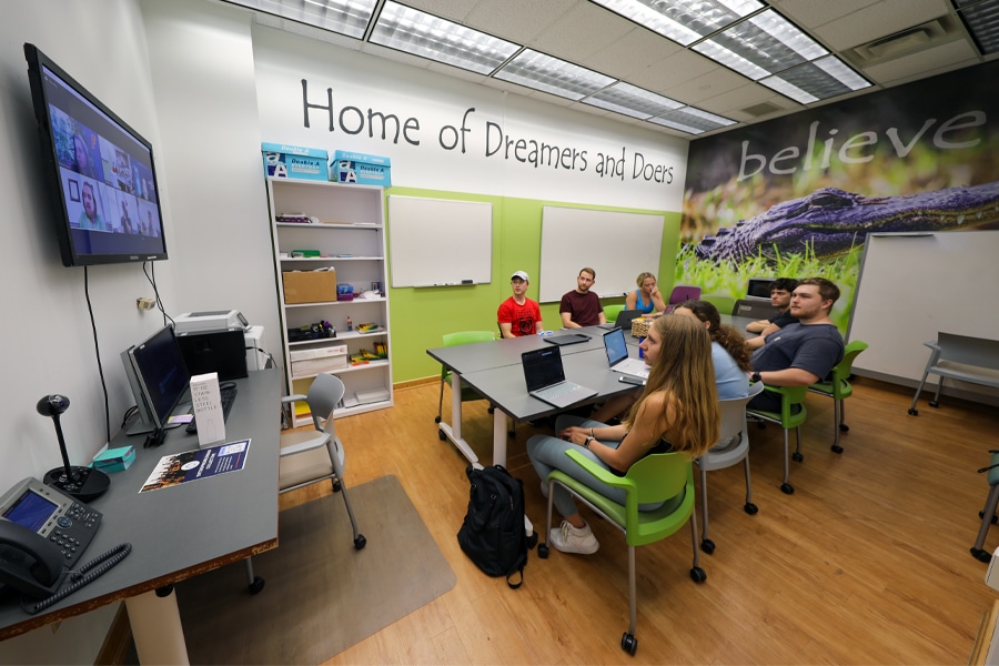 A group of students in a brightly lit classroom, seated around tables with laptops, listening attentively to a virtual meeting displayed on a screen. Inspirational words like “believe” and “Home of Dreamers and Doers” are painted on the walls.