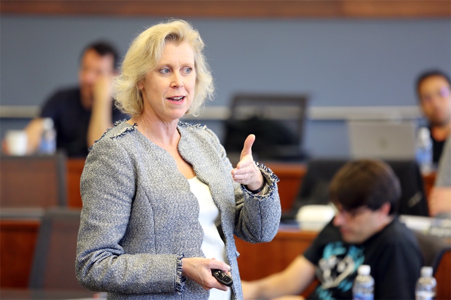 A professional woman with blonde hair wearing a light gray blazer gestures while speaking in a classroom setting. In the background, several students are seated and engaged.