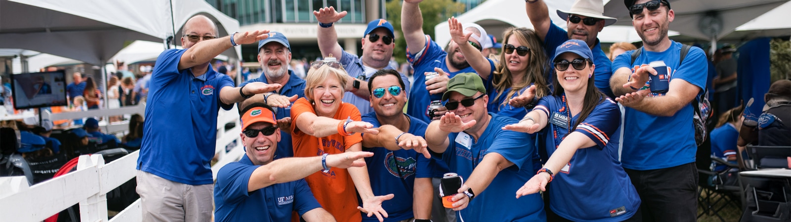 A lively group of people wearing blue and orange Florida Gators apparel pose together outside under tents, smiling and enthusiastically reaching their hands forward in a celebratory gesture.