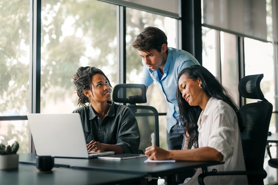 Three professionals collaborating at a desk with a laptop in a bright office space. One woman is typing on the laptop, another is taking notes, and a man is leaning over to discuss.