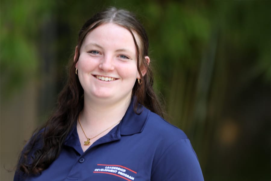 A young woman with long, wavy brown hair smiles at the camera, wearing a navy blue Leadership Development Program polo shirt, with a soft outdoor background.