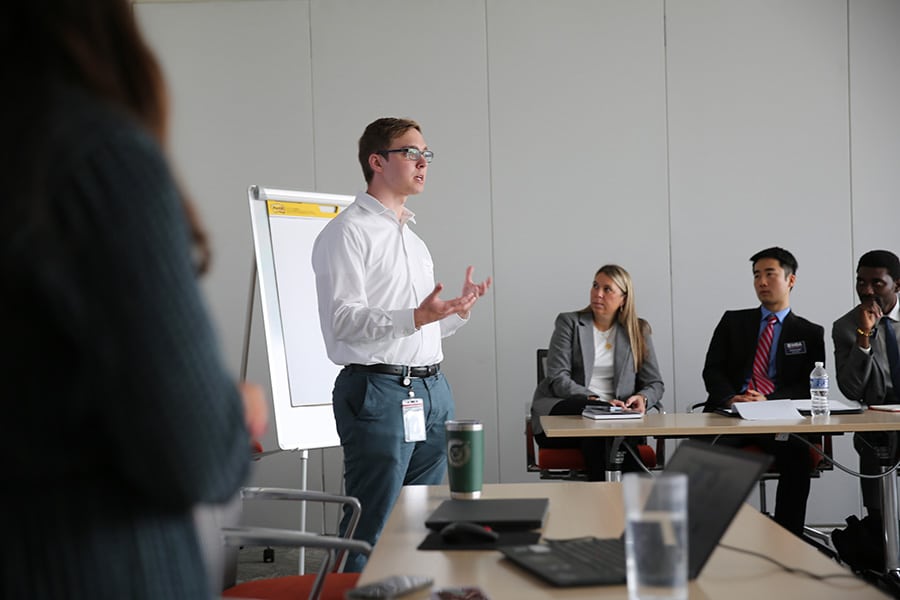 A man standing and presenting in front of people around him seated at tables.