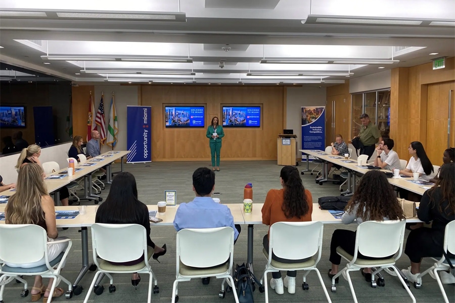 A group of graduate students sit around a U-shaped table, listening to a speaker in a modern classroom with presentation screens displaying a welcome message for a marketing consulting course.