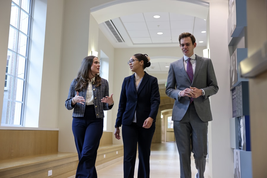 Three graduate students in business attire walk and talk together in a bright, modern hallway lined with windows and portraits.
