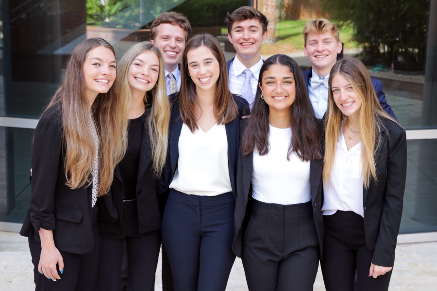 A group of nine business students, dressed professionally in blazers and shirts, smile brightly as they stand closely together outside a modern building.