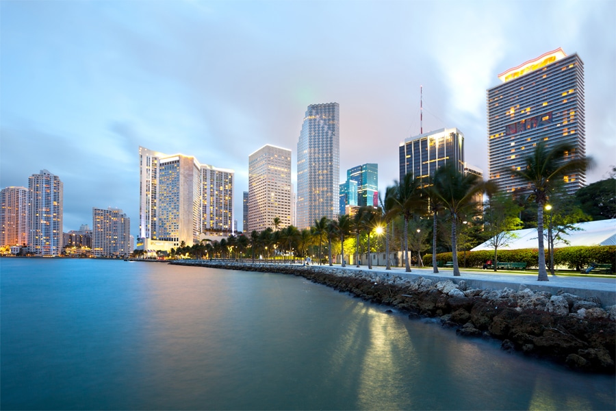 View of downtown Miami skyline at dusk with tall buildings, palm trees along the waterfront, and calm water in the foreground.