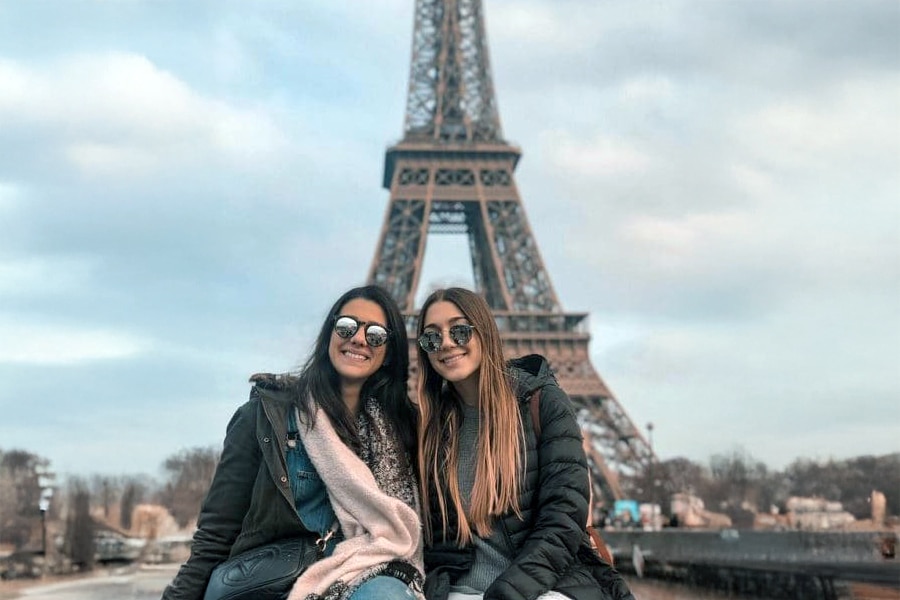 Two smiling young women wearing winter coats and sunglasses sit closely together in front of the Eiffel Tower in Paris, France, with a cloudy sky overhead.