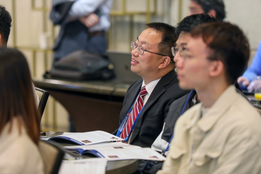 A man in a suit and red-striped tie, wearing glasses and a Warrington lanyard, sits attentively among other attendees at a professional conference, looking engaged.