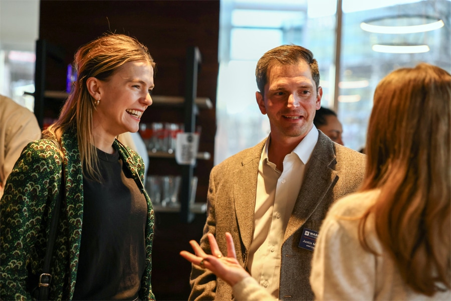 Professionally dressed individuals engaged in conversation during a networking event, with several others mingling in the background of a well-lit conference room.