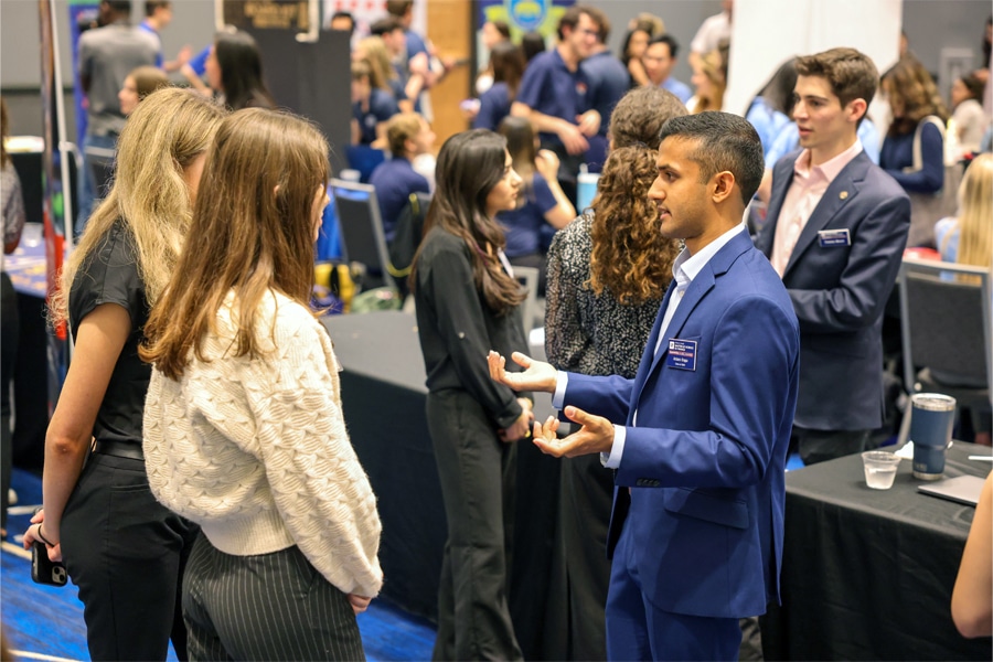 A well-dressed young man in a blue suit engages in conversation with three young women at a busy event filled with students and professionals networking.