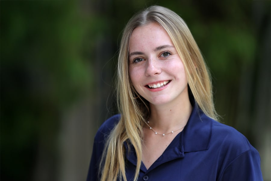 A young woman with straight blonde hair smiles brightly at the camera, dressed in a navy blue Leadership Development Program polo shirt, with a natural outdoor background.