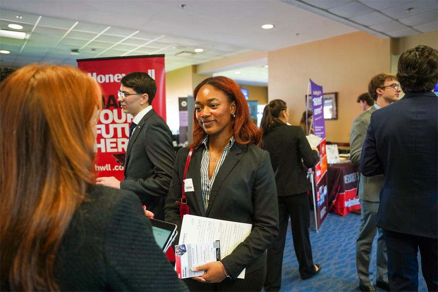 A young woman dressed in business attire holds a résumé and informational pamphlets while engaging in conversation with a recruiter at a career fair. Other students and recruiters are visible in the background.