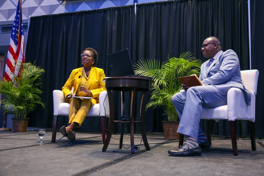 Two speakers on a stage, a woman in a yellow suit and a man in a light blue suit, engaged in discussion with a laptop, notepads, and a small table between them.