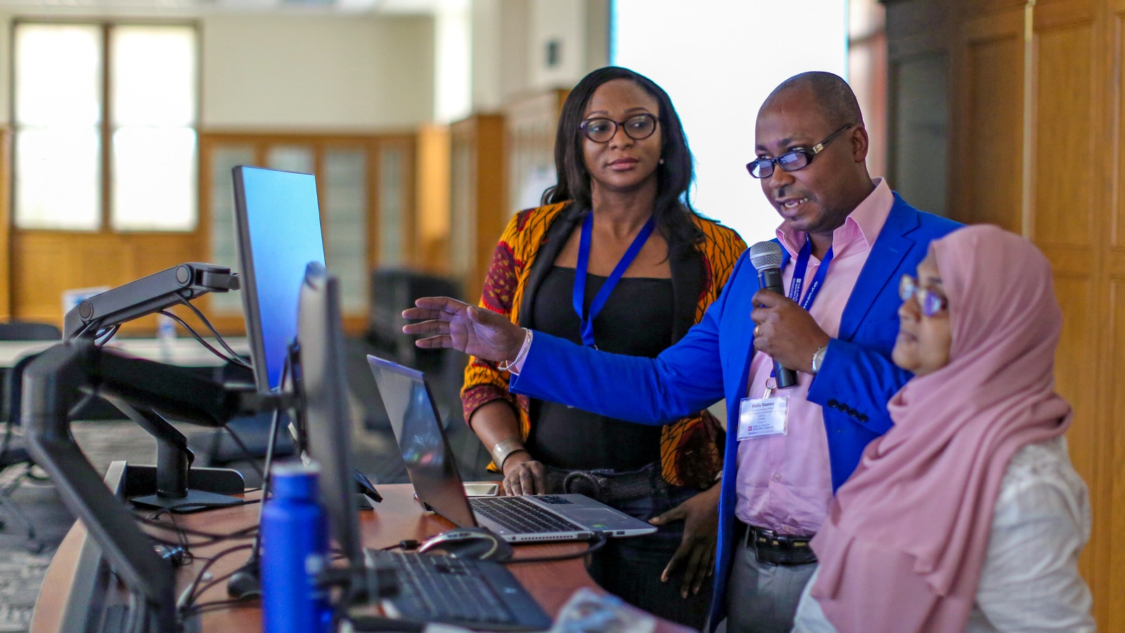 Three professionals — one man in a bright blue blazer holding a microphone and two women, one in traditional attire — present a project using multiple computer monitors in a classroom-like setting.