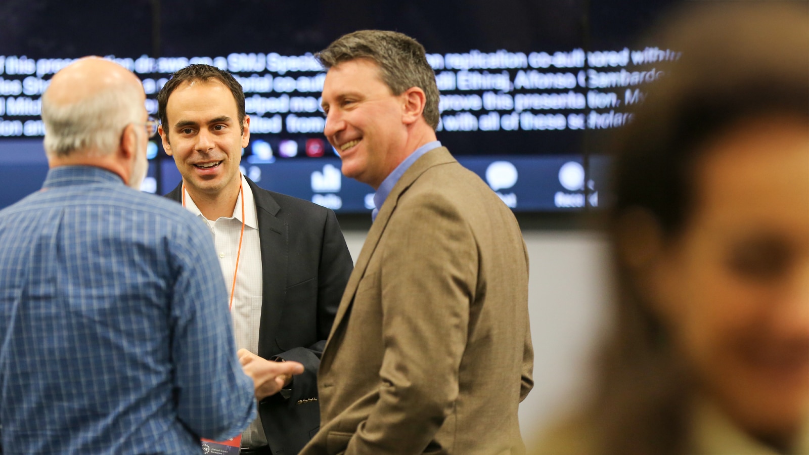 Three professionals network and converse at a business event, smiling and engaging in discussion, with a presentation screen in the background.
