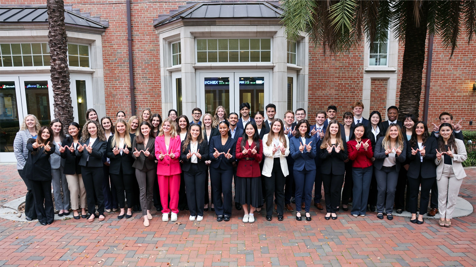 A large group of professional-looking students stand outside a brick building at the University of Florida, smiling and holding up “W” hand signs to represent Warrington.