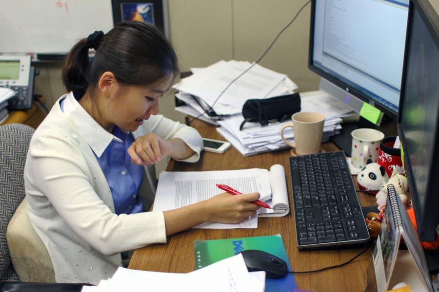 A Ph.D. student sits at a desk covered in papers, notes, and mugs, intently marking up a research paper with a red pen while working between a large monitor and open books.