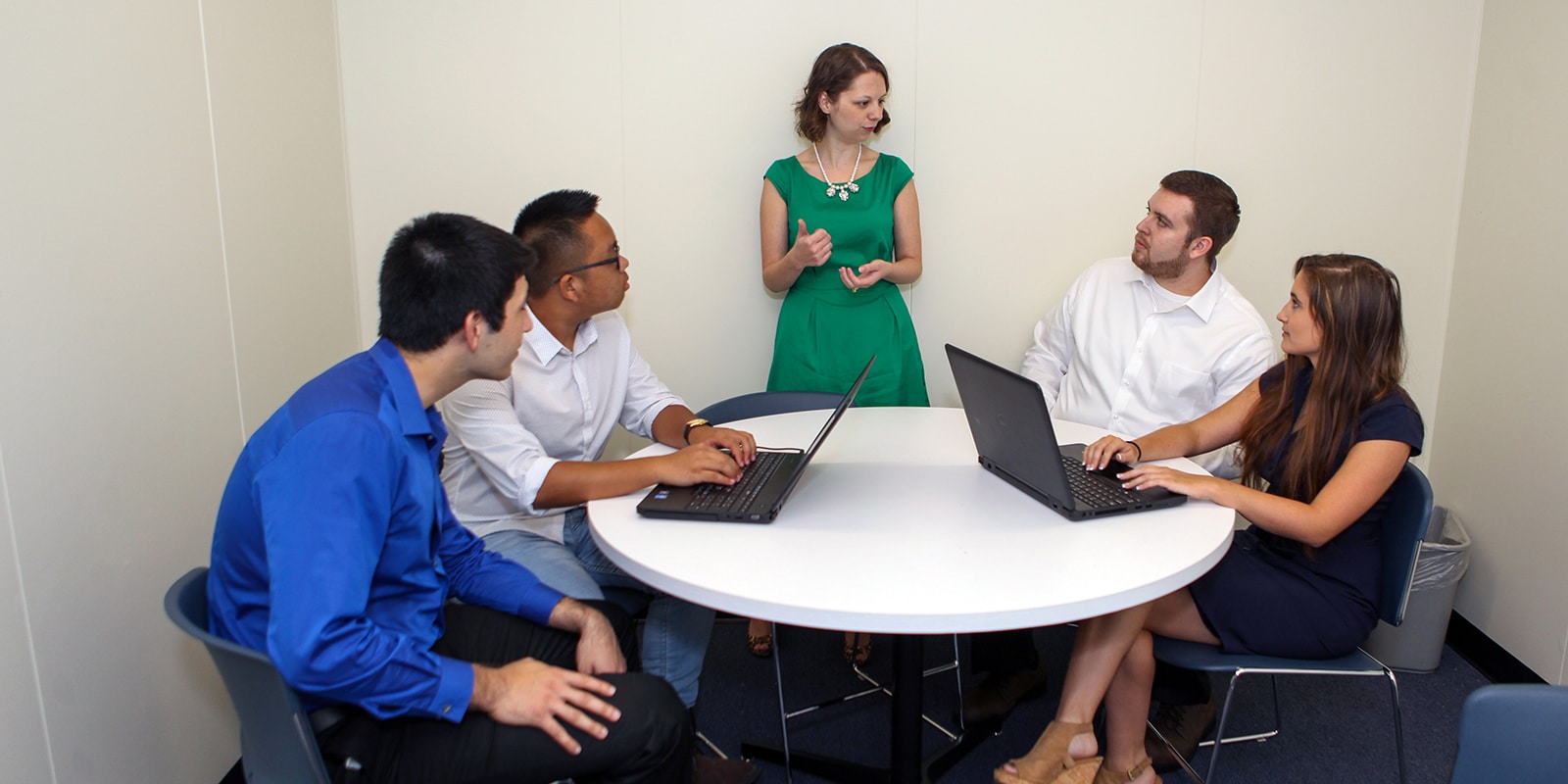 Standing, a UF Warrington faculty member speaks to a group of PhD in Finance and Real Estate students, sitting at a table with laptops, about their dissertations