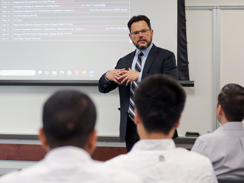 Professor Murillo Campello lectures to Ph.D. students in a business classroom at the University of Florida, with a syllabus projected on the screen behind him.