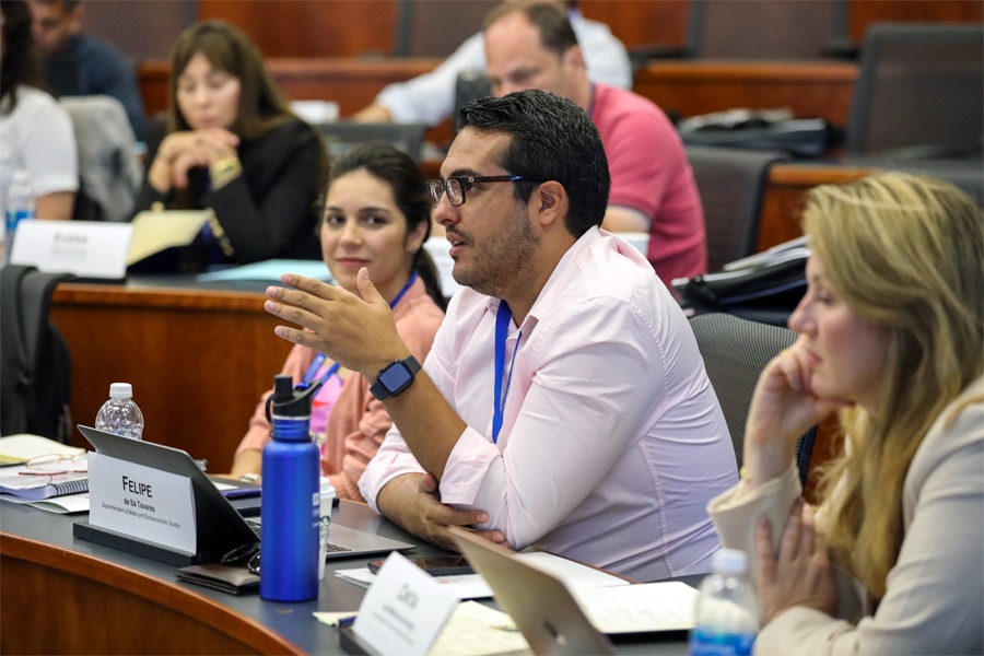 Graduate students and faculty engaged in discussion in a tiered classroom. A man wearing glasses and a light pink shirt is speaking while gesturing with his hand.