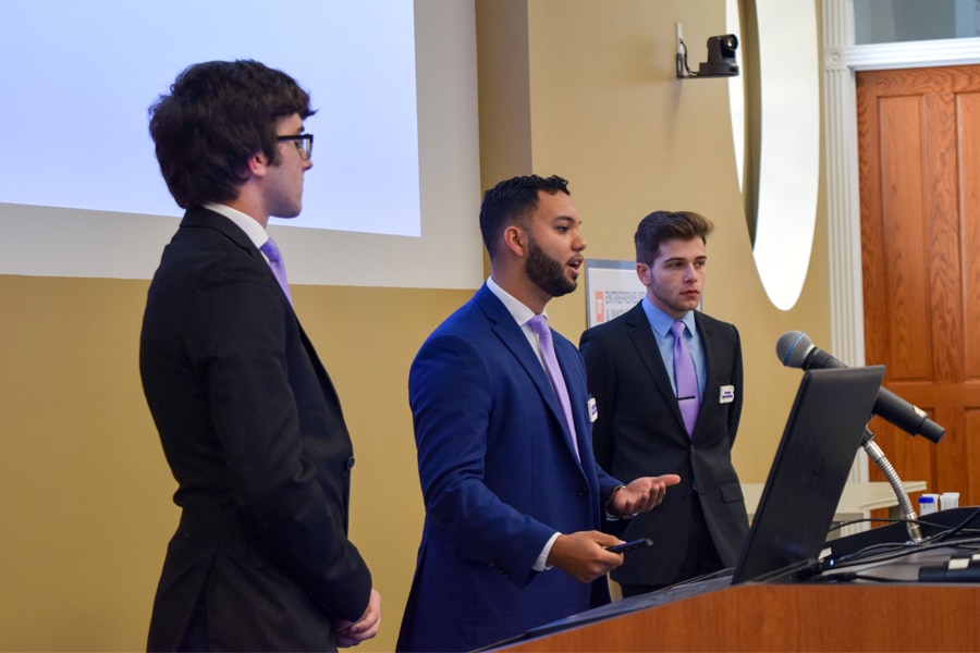 Three well-dressed student competitors standing at a podium in a classroom, presenting their business plan to an audience.